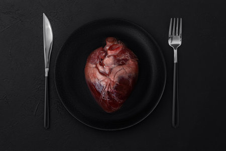 Raw red heart in a black plate with fork and knife on a black background