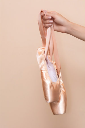 A woman's hand holds pink ballet flats on a beige background. Pointe shoes