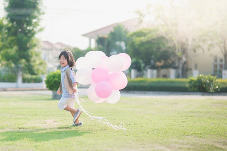 Cute Asian child with many balloons playing in the park under sunlight on summer dayの写真素材