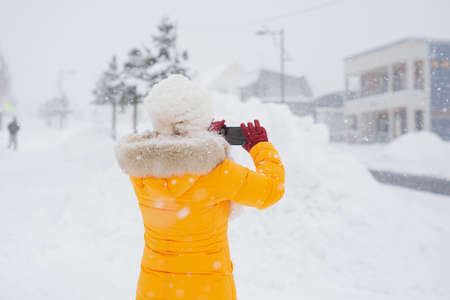 Asian woman wearing yellow clothes taking photo with mobile phone on city street during snowfall in winter.の写真素材