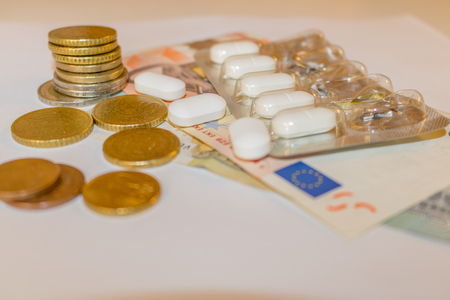 White Tabletts with euros banknotes and coins. Concept Pharmaceutical business, cost of health, medecine. Scattered pills.の写真素材