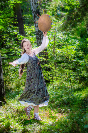 Young woman of a sorceress performs a ritual dance with a tambourine in the forest on a summer sunny day.の写真素材