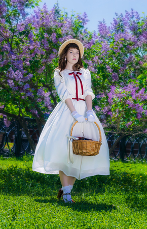 girl in a white dress with a basket in her hands in a walk in the garden on the background of a bush of blossoming lilacs.の写真素材