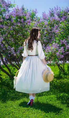 Woman in a white dress with a basket in her hands in a walk in the garden on the background of a bush of blossoming lilacs.の写真素材