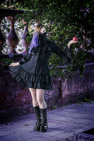 Woman in a black dress and mask with an apple in her hands.