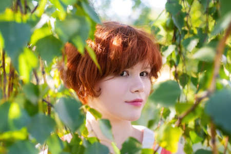 Portrait of a red-haired happy woman in park among the green foliage.
