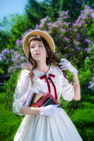 Woman in a white dress with a books in her hands in on a walk in the garden against the background of a bush of blossoming lilacs.の写真素材