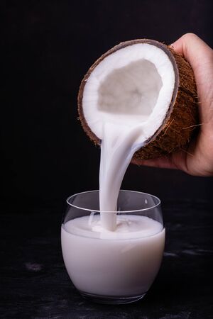 Front View, coconut milk contained in a half coconut is poured into the glass on a dark backgroundの写真素材
