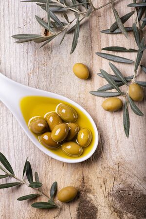 High angle view, white ceramic bowl, with extra virgin olive oil and green olives, on rustic wooden table with olive branchの写真素材
