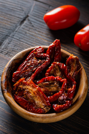 On the dark wooden table, a wooden bowl with dried tomatoes softened with olive oil. A few fresh tomatoes aside.の写真素材