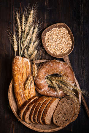 on the rustic wooden table, a wooden plate with different types of bread, with various seeds and cereals and a few ears of wheat. Still life. view from top.の写真素材