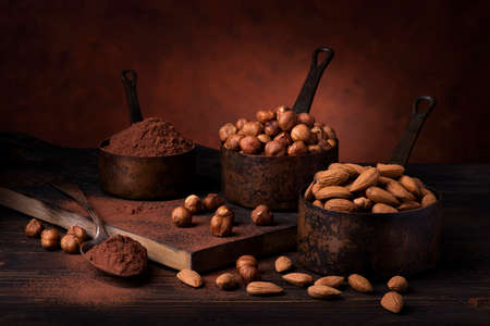 Still life. in the foreground with dark background, vintage metal measuring spoons with shelled hazelnuts and almonds and cocoa powderの写真素材