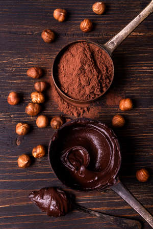 In the foreground, top view, on a rough wooden background, vintage metal measuring spoons with melted dark chocolate and cocoa powder with shelled hazelnutsの写真素材