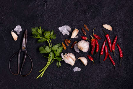 a bunch of fresh parsley, a metal scissor, cloves of garlic and hot peppers in the foreground, on a textured black background. View from aboveの写真素材