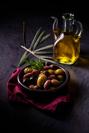 in the foreground a wooden bowl with seasoned and spiced olives. In the background a glass cruet with extra virgin olive oil. Still lifeの写真素材