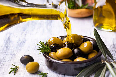 in the foreground, extra virgin olive oil is poured into a bowl with rosemary-flavored olivesの写真素材