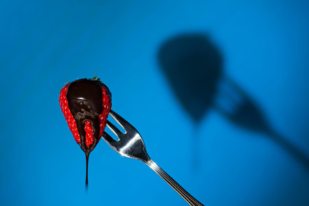 Close up of a strawberry suspended from a fork and its shadow on background with liquid chocolate aboveの写真素材
