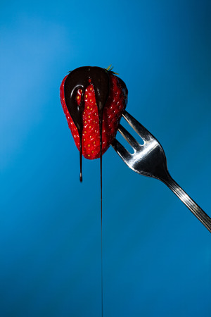 Close up of a strawberry suspended from a fork with liquid chocolate above on a blue backgroundの写真素材