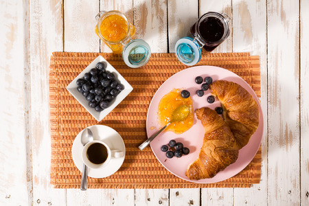 Breakfast with croissant, blueberries, coffee, orange and blueberry jam over a tablecloth seen from aboveの写真素材