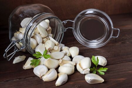 foreground of an open glass airtight jar with some spilled garlic cloves and parsley leaves on a table and wooden backgroundの写真素材