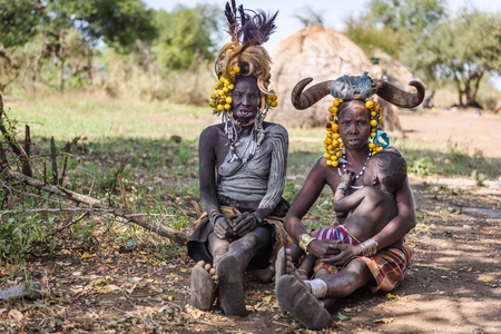 MAGO NATIONAL PARK, ETHIOPIA - AUGUST 19, 2015:  unidentified Mursi women and child with  beautiful fruit decoration on their head and animal horns. The old one has the lips open to insert a lip plateのeditorial素材