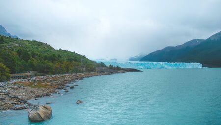 blue lake with rocks and a walkway on the coast surrounded by trees and mountains with the Perito Moreno glacier in the background under a cloudy skyの写真素材