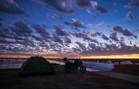 tent and motorcycle on the coast of a river next to a bridge at dawnの写真素材