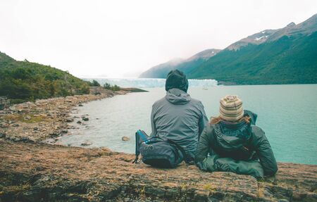 couple of tourists resting sitting on a rock on the shore of a blue lake surrounded by trees and mountains with the Perito Moreno glacier in the background under a cloudy skyの写真素材