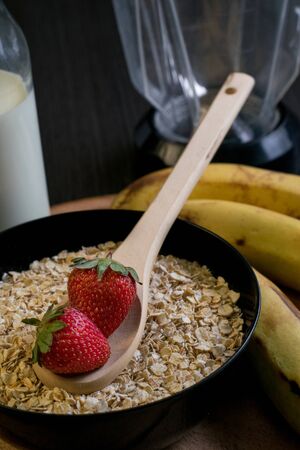Close-up of two strawberries in a wooden spoon on a bowl full of oatmeal with bananas and a bottle of milk in the backgroundの写真素材