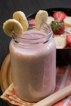 Banana, strawberry and oatmeal smoothie in a jar with slices of bananas and strawberries in a bowl full of oatmeal and next to a wooden spoon with oatmeal, top of a rag with a round wooden downの写真素材