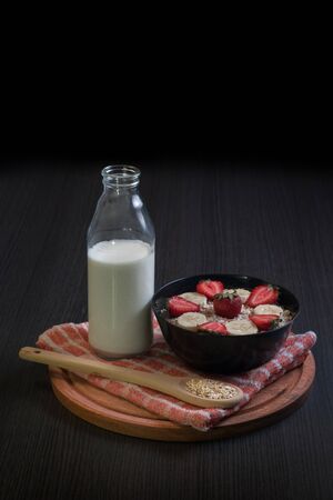 general view of a bowl of oatmeal with a wooden spoon with two strawberries with a bottle of milk and bananas around and a jug of blender on top of a round wooden boardの写真素材