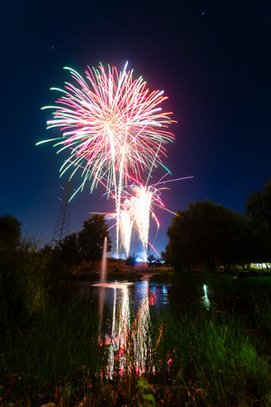 Bright fireworks burst in vibrant colors above a serene pond, reflecting lights on the water's surface at night.の写真素材