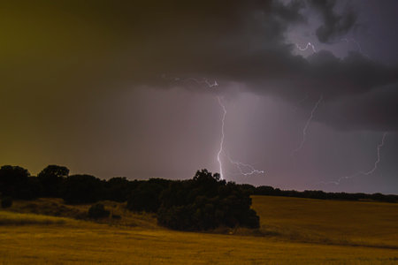 Dark clouds gather as lightning strikes, creating a dramatic display in the night sky above a rural landscape.の写真素材