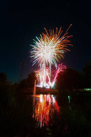 Vibrant fireworks burst in various colors over a calm lake, reflecting the light and beauty during a summer night.の写真素材