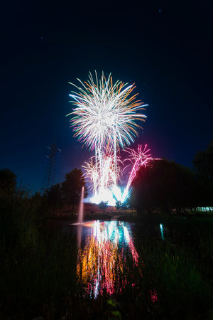 Bright fireworks burst in vibrant colors above a calm pond, reflecting their beauty on a warm summer...
