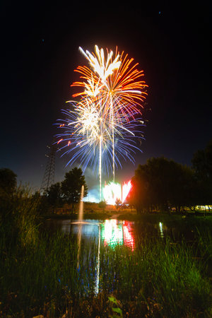 Bright fireworks burst in various colors over a calm pond, reflecting vividly in the water at night.の写真素材