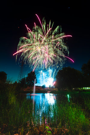 Bright fireworks illuminate the night sky above a tranquil pond surrounded by trees and greenery.の写真素材