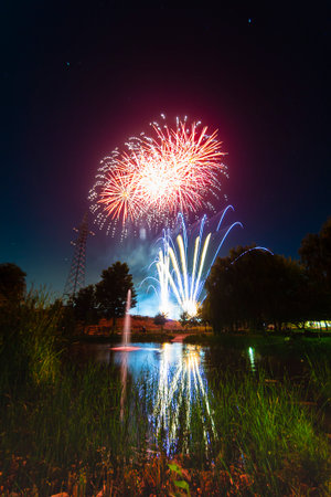 Colorful fireworks burst above a serene pond, creating bright reflections on the water during a summer evening.の写真素材