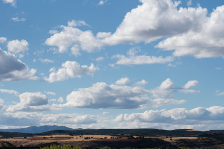 A vast sky filled with fluffy white clouds overlooks gentle hills and fields under sunlight in nature's tranquility.の写真素材