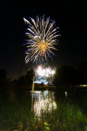Bright fireworks light up the night sky, reflecting in a calm lake amidst lush greenery and trees.の写真素材