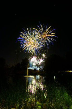 Brightly colored fireworks burst in the night sky above a serene lake, creating reflections in the water.の写真素材