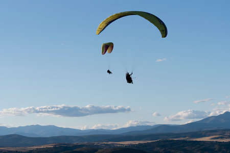 Two paragliders gracefully descend against a backdrop of expansive mountains and a bright blue sky.の写真素材