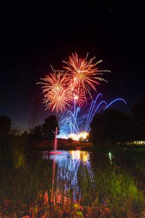 Bright fireworks burst in vibrant colors above a calm lake, reflecting on the water during a summer...