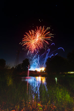 Bright fireworks burst in the sky, creating colorful reflections on a calm lake, surrounded by trees...