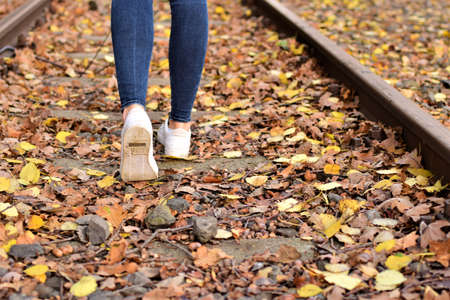 Walking legs wearing blue jeans and white shoes on autumn leaves and old railroad tracksの写真素材