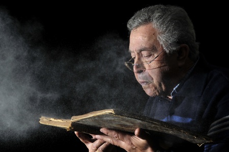 Senior man blowing dust of  a very old book isolated in blackの写真素材
