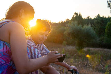 Young woman showing a video or photo in the phone to a boy in a bicycle at sunset outdoorsの写真素材