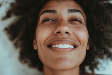 Radiant close-up of a joyful woman with a beaming smile, her eyes closed in happiness, and natural curly hair framing her faceの素材