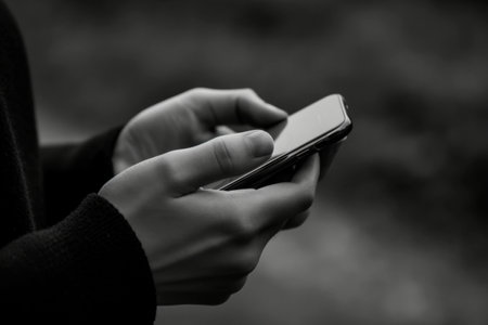 Monochrome image of a person's hands texting on a smartphone, set against a blurred natural backgroundの素材