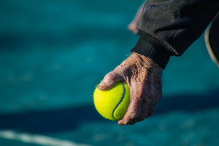 Elderly hand gripping a tennis ball against a blurred court background, symbolizing active agingの素材